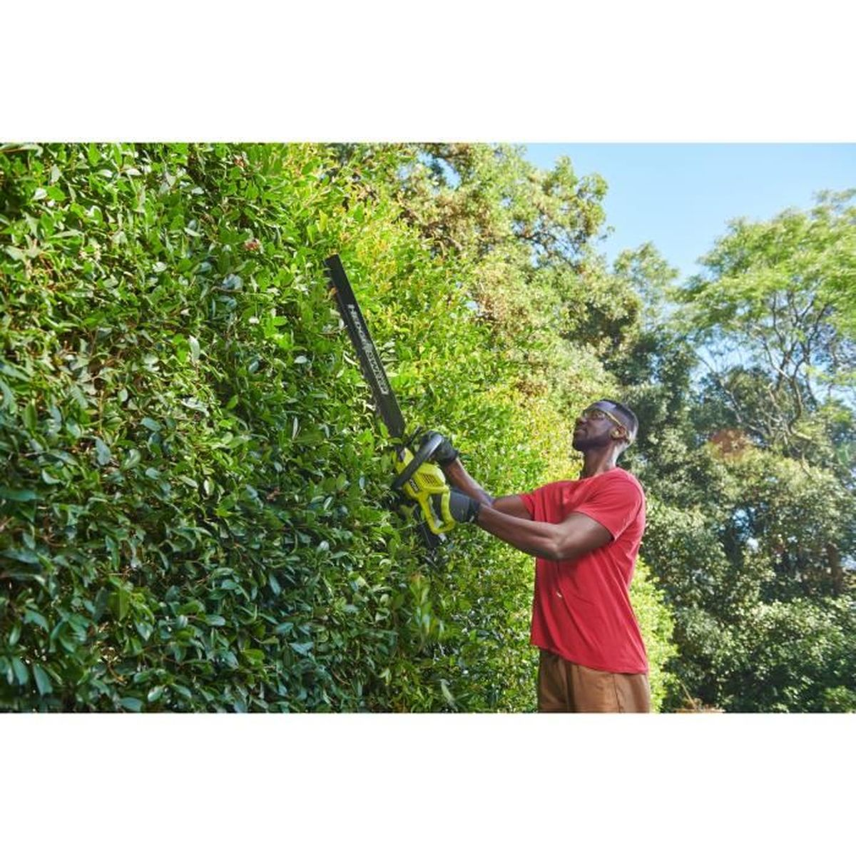 Ein Mann im roten Hemd schneidet draußen unter Bäumen eine hohe grüne Hecke mit einer Heckenschere.