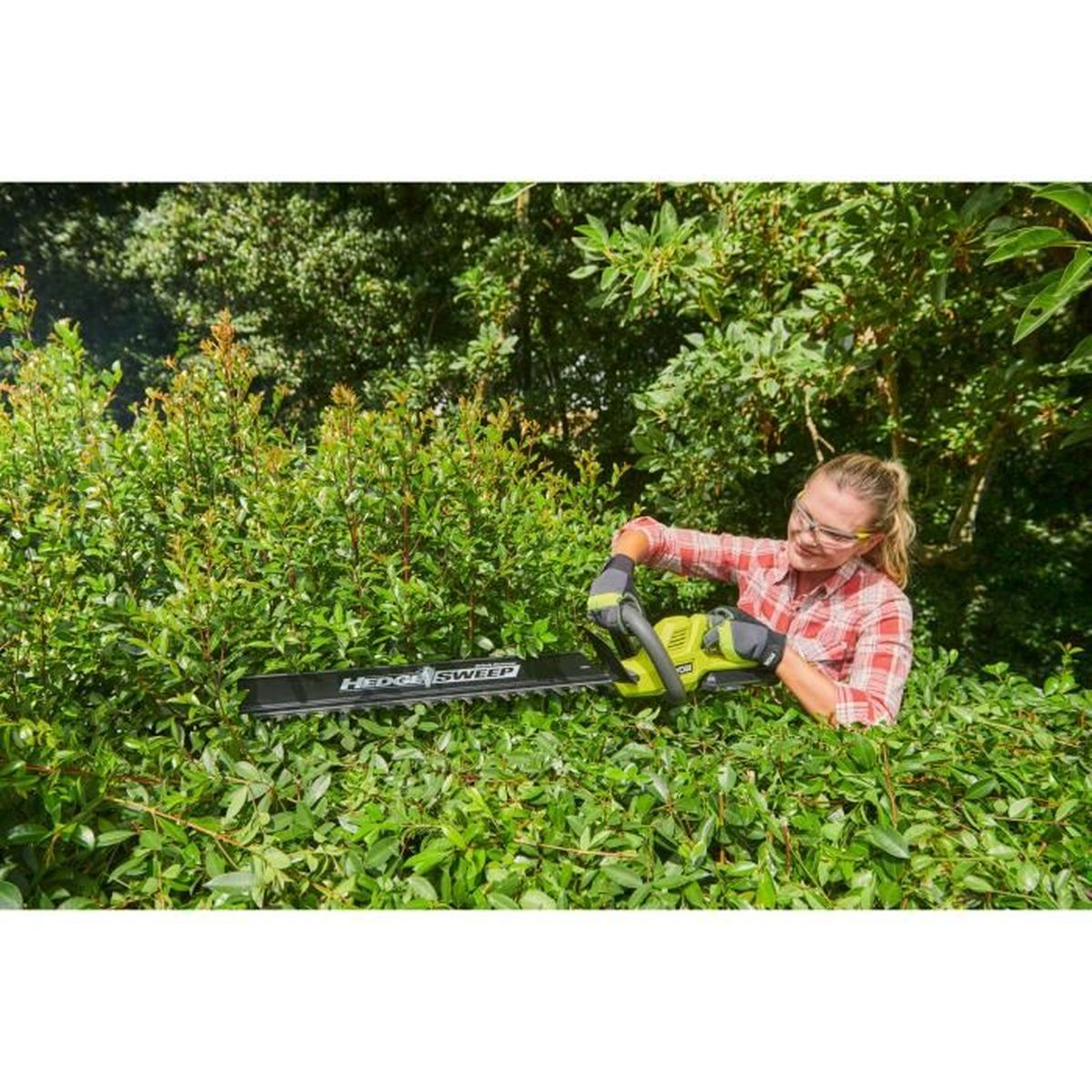 Eine lächelnde Frau schneidet in einem üppigen Garten eine grüne Hecke mit einer Akku-Heckenschere.