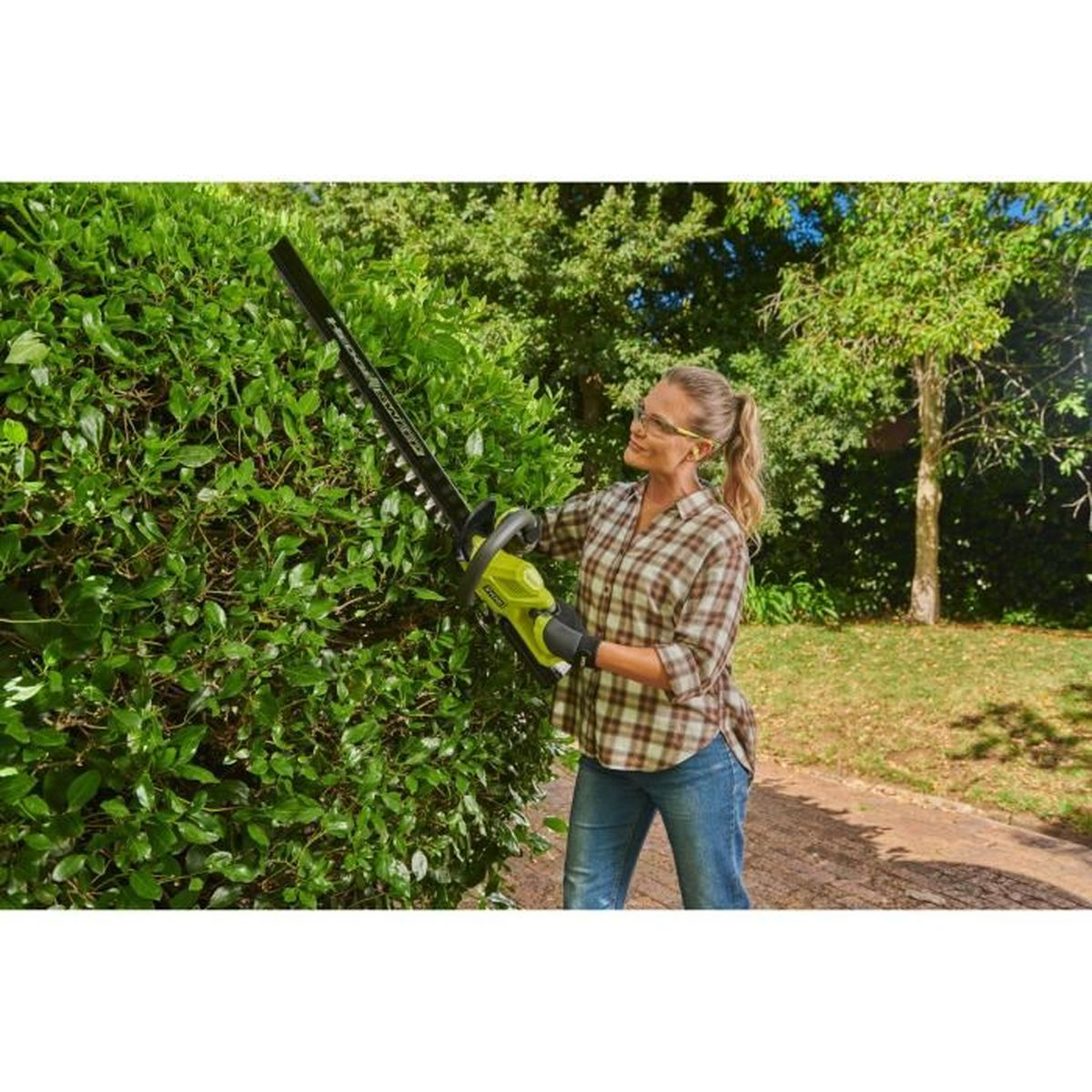 Eine Frau schneidet mit einer elektrischen Heckenschere einen grünen Busch in einem Garten.