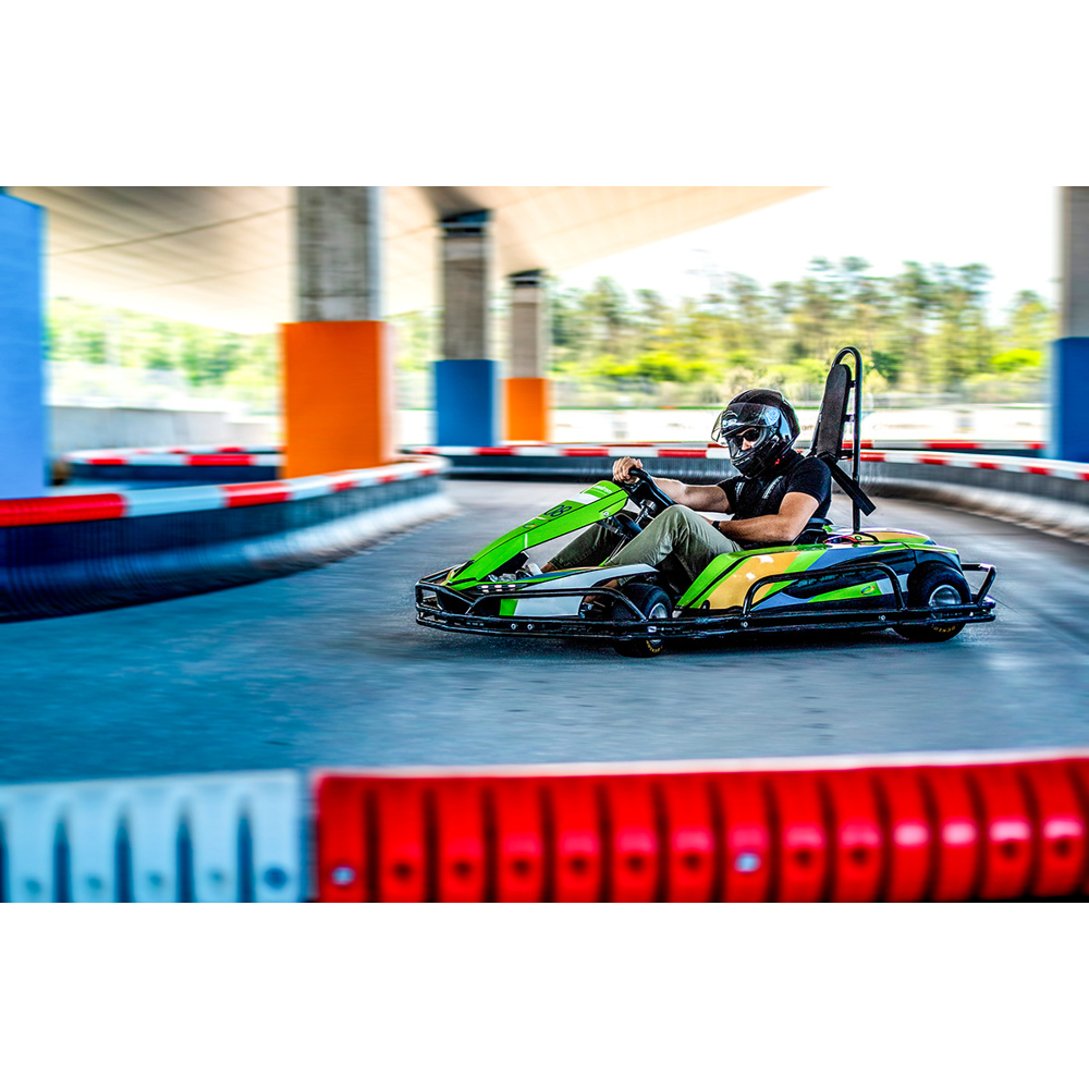Ein Fahrer mit Helm fährt in einem grünen Go-Kart über eine Indoor-Rennstrecke mit rot-weißen Barrieren.