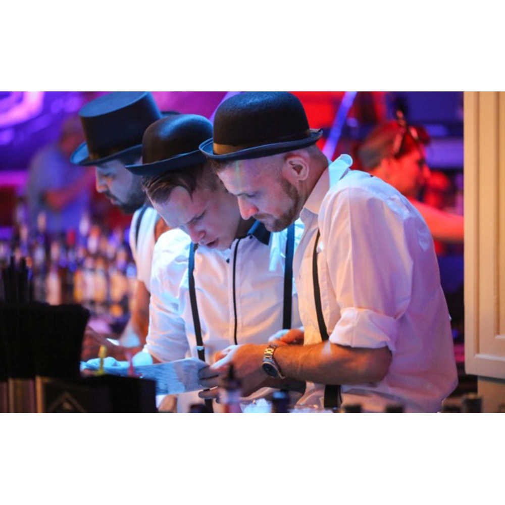 Drei Barkeeper in weißen Hemden, Hosenträgern und Hüten schauen gemeinsam auf einen Zettel hinter der Bar.
