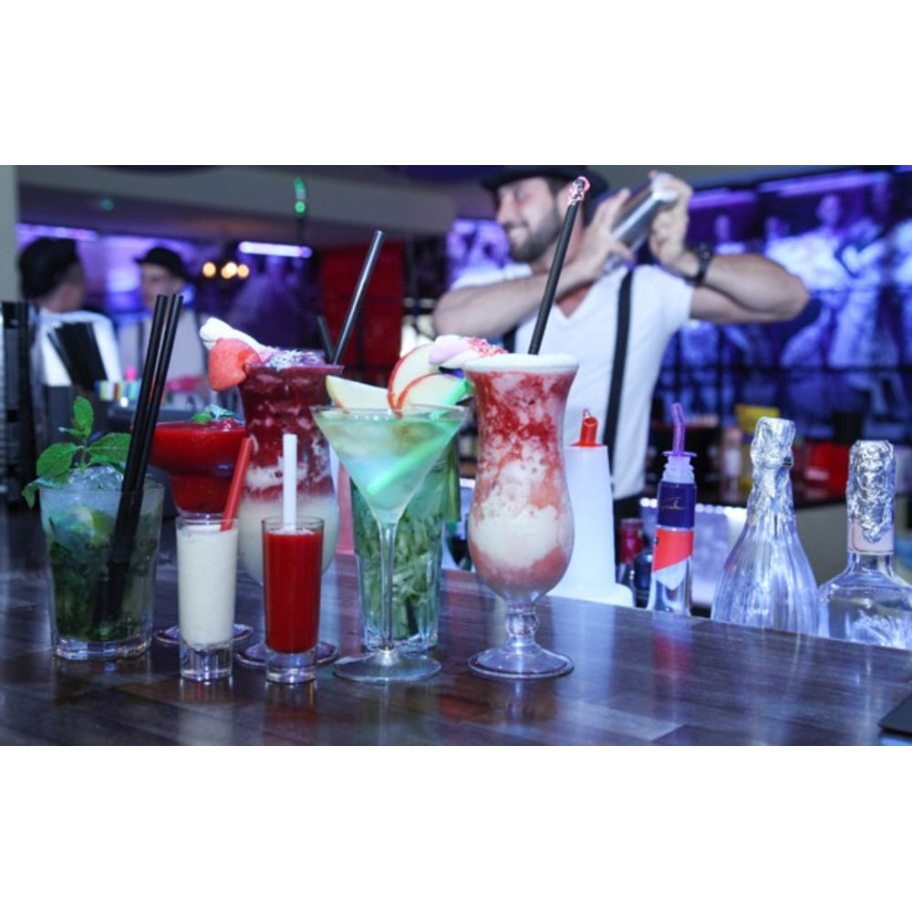 Bunte Cocktails stehen an einer Bar, im Hintergrund schüttelt ein Barkeeper einen Shaker.