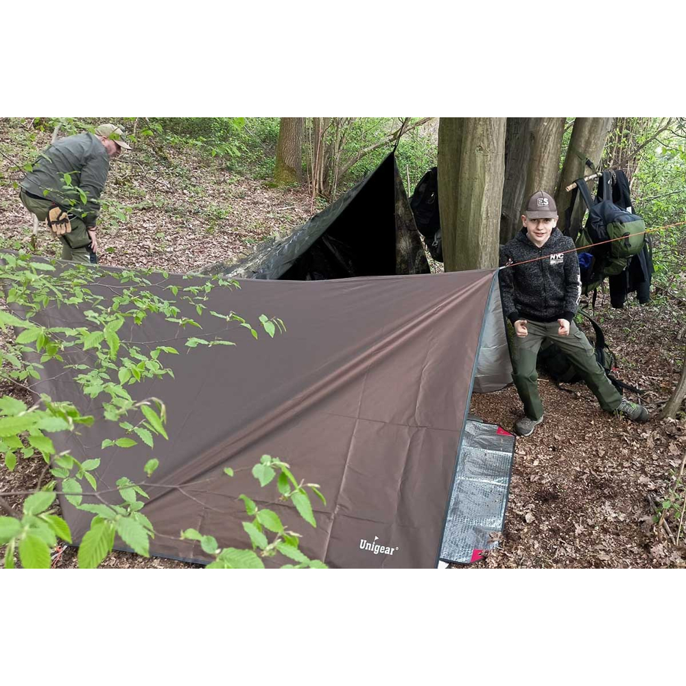 Zwei Personen im Wald beim Aufbau eines braunen Unigear-Tarps, im Hintergrund hängen Rucksäcke an Bäumen.
