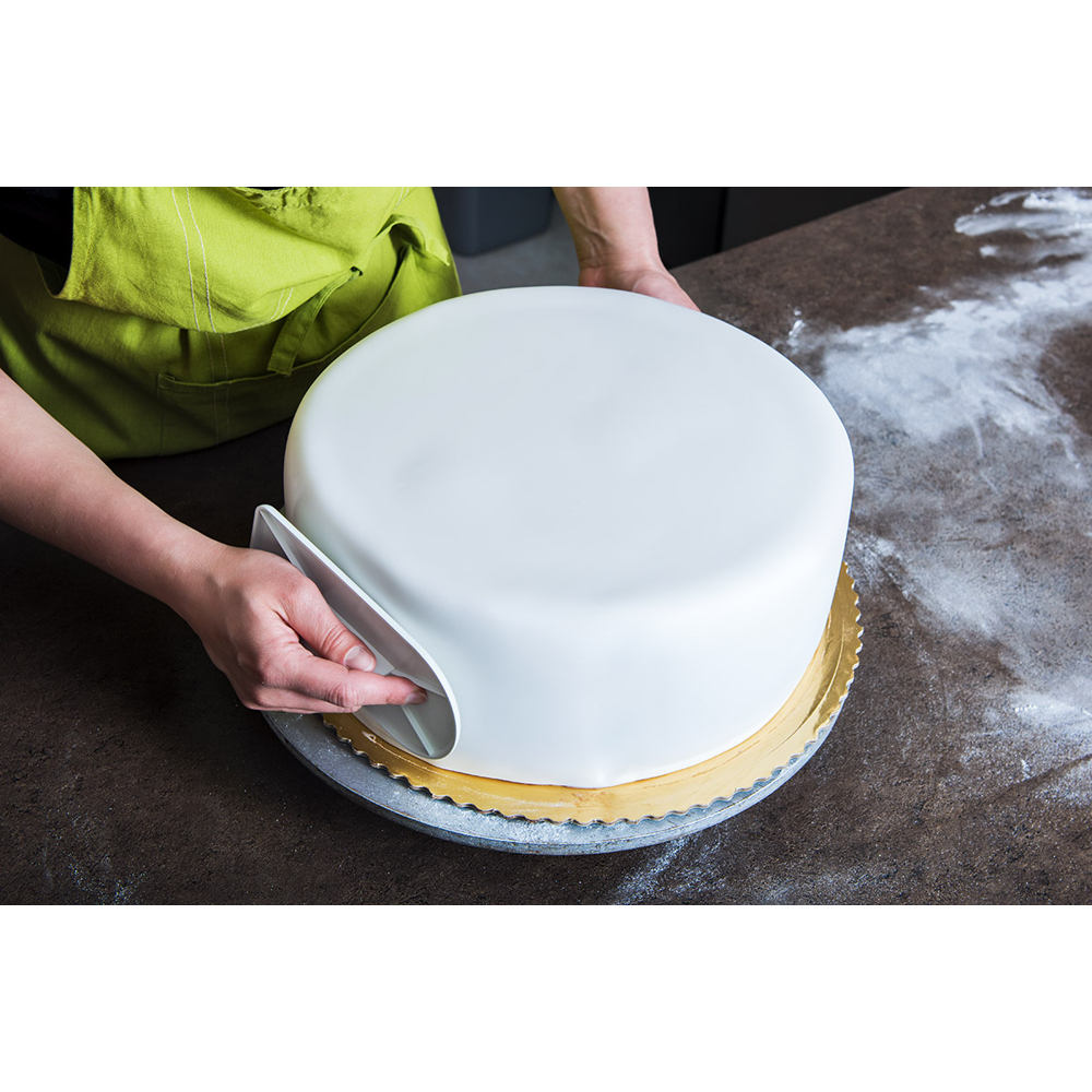 Eine Person glättet mit einem weißen Schaber weißen Fondant auf einer runden Torte.
