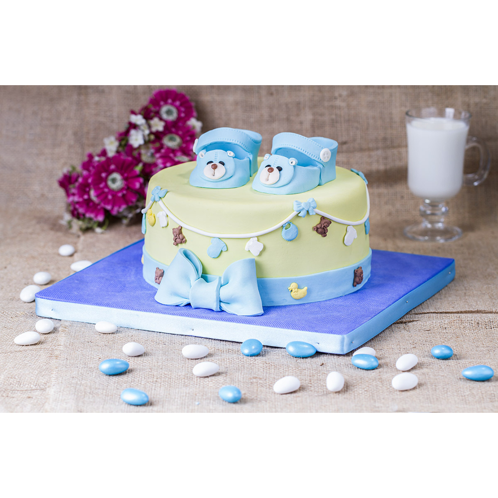 Eine hellgrüne Babytorte mit blauen Schuhen, einer großen Schleife, Blüten im Hintergrund und Bonbons.