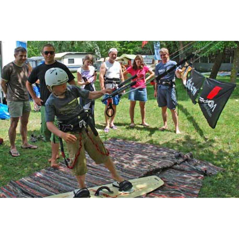 Ein Kind mit Helm und Kitesurf-Ausstattung übt auf einer Matte im Gras, beobachtet von Erwachsenen.