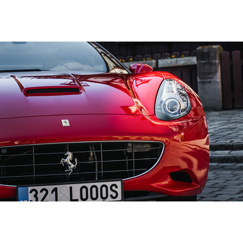 Nahaufnahme eines roten Ferrari Sportwagens auf kopfgepflasterter Straße vor einem Holzzaun.