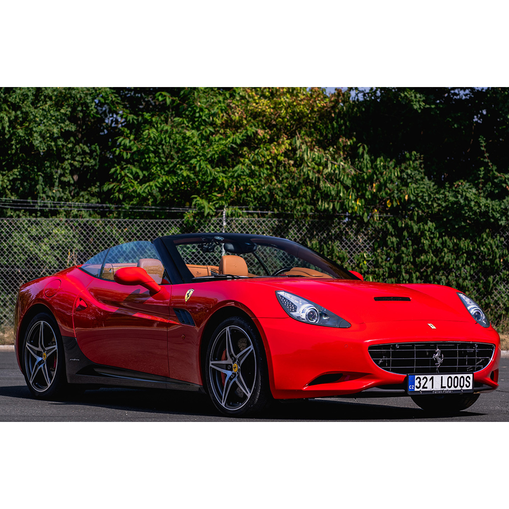 Ein rotes Cabriolet-Sportauto, geparkt auf Asphalt vor grünen Bäumen und einem Maschendrahtzaun.
