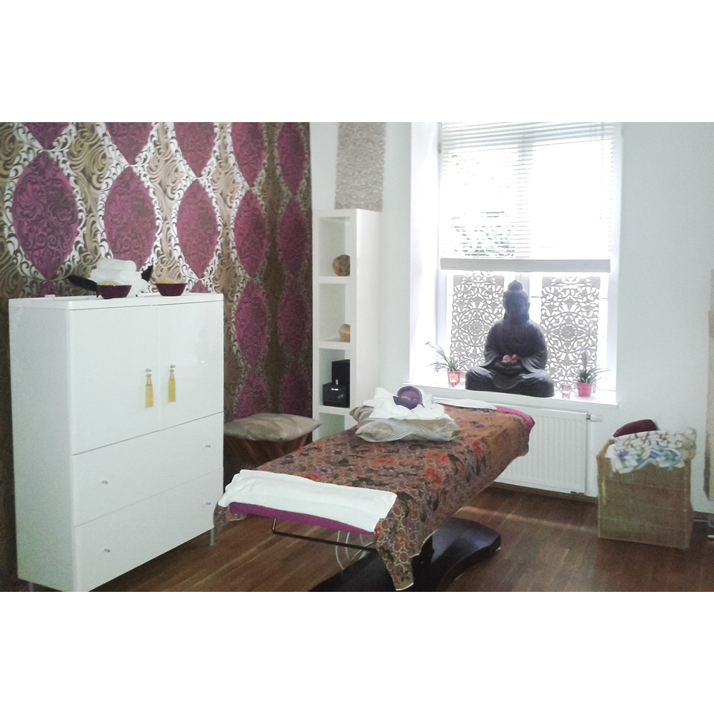 Ein heller Massageraum mit einer Liege, einem weißen Schrank, einer Buddha-Statue und dekorativen lilafarbenen Tapeten.