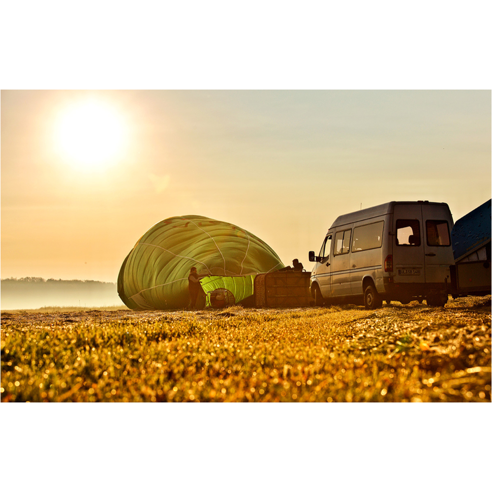 Ein gelber Heißluftballon wird bei Sonnenaufgang neben einem Transporter auf einem Grasfeld vorbereitet.