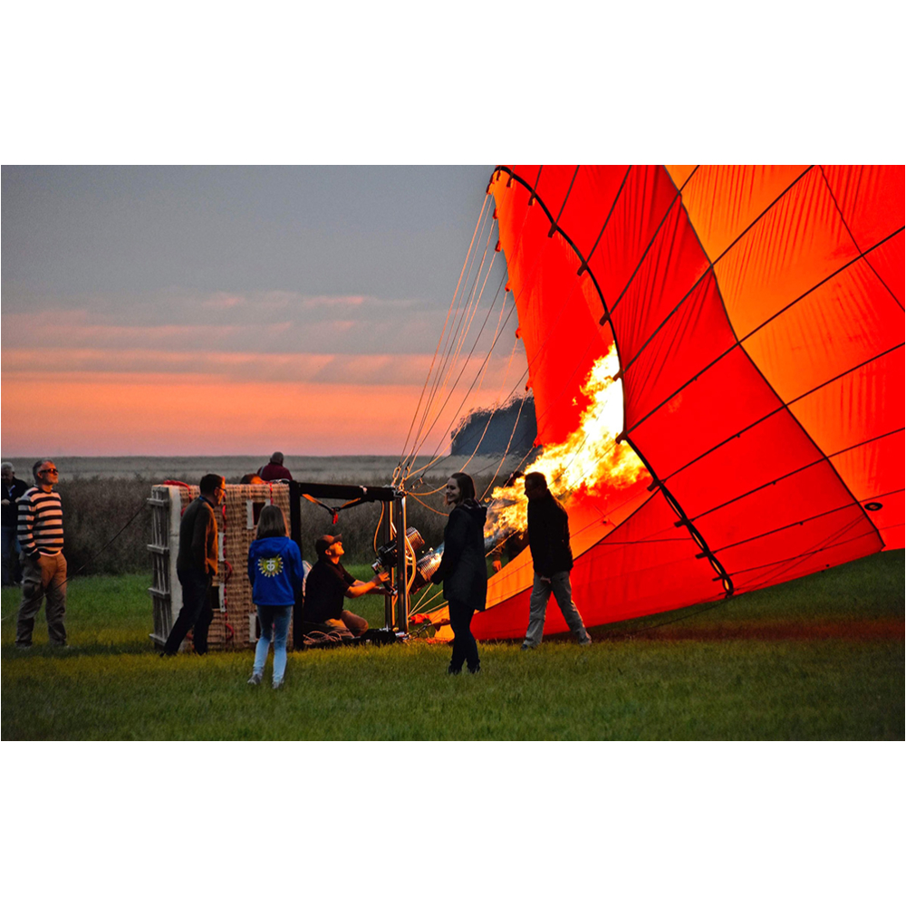 Menschen versammeln sich in der Dämmerung auf einer Wiese, während ein orangefarbener Heißluftballon befeuert wird.