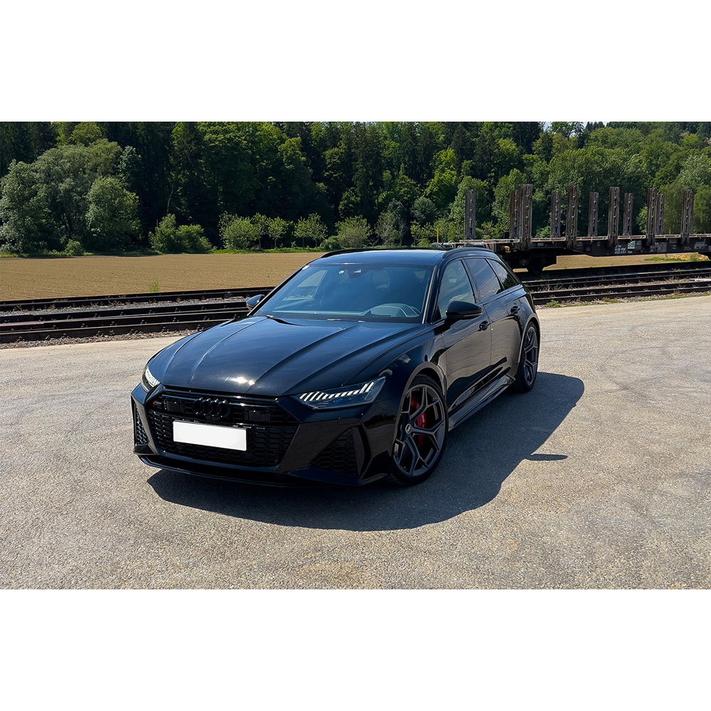 Ein schwarzer Audi RS6 Kombi parkt auf einem asphaltierten Platz vor Bahngleisen und einem grünen Wald.