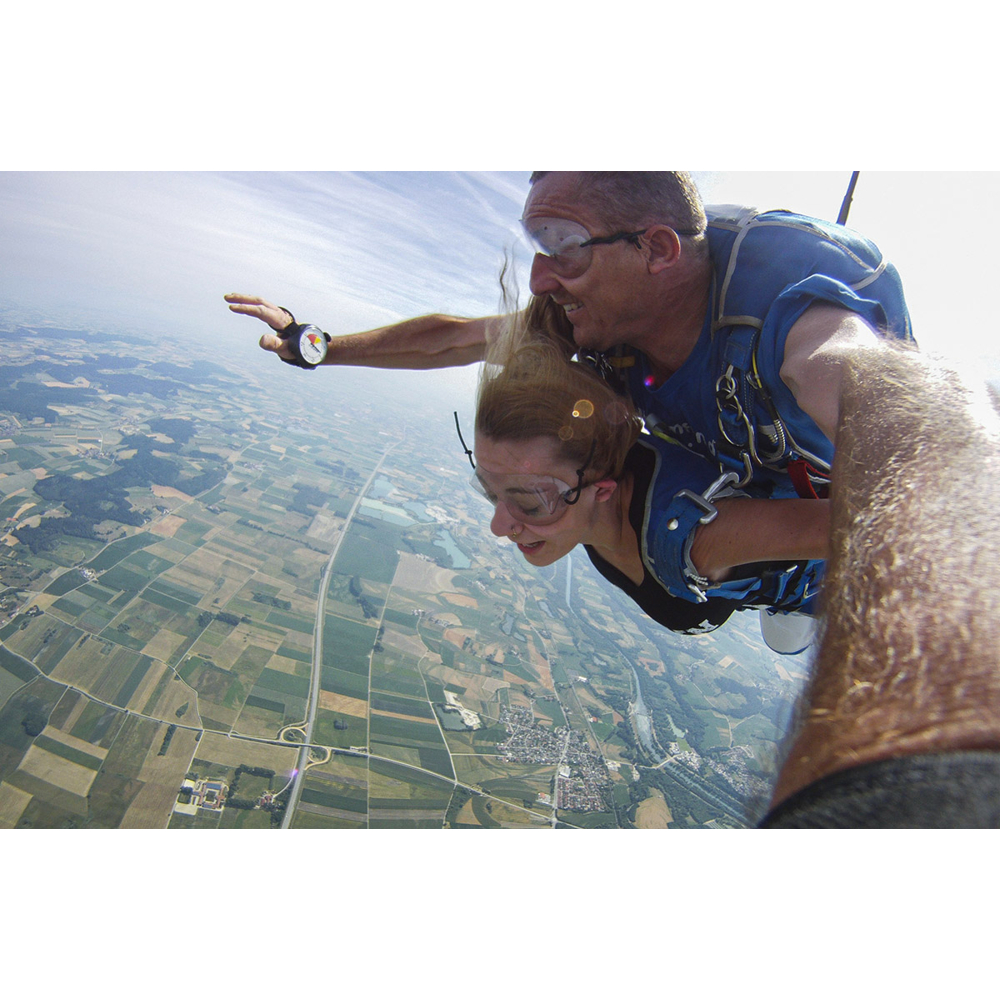 Tandem-Fallschirmspringer im Flug über einer grünen Landschaft mit Feldern und Straßen.