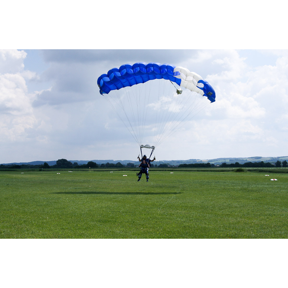 Zwei Fallschirmspringer in blauen und weißen Schirmen landen auf einer grünen Wiese unter wolkigem Himmel.