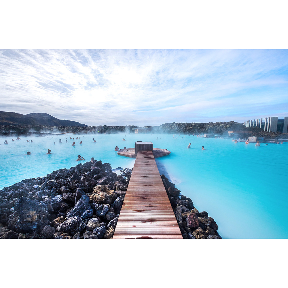 Holzsteg führt in milchig-blaue Thermalquelle mit vielen Badegästen vor Vulkanlandschaft.