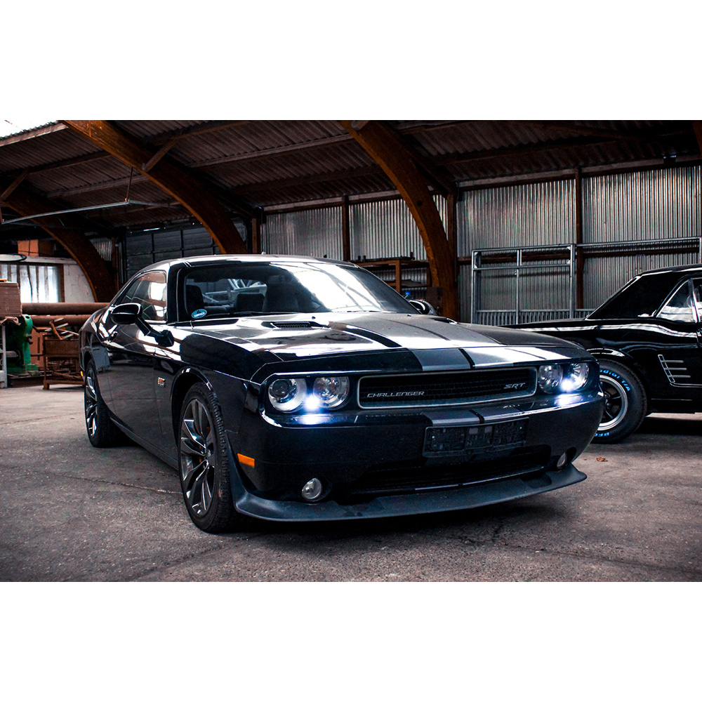 Ein schwarzer Dodge Challenger SRT mit eingeschalteten Scheinwerfern steht in einer großen Industriehalle mit Holzbalken.