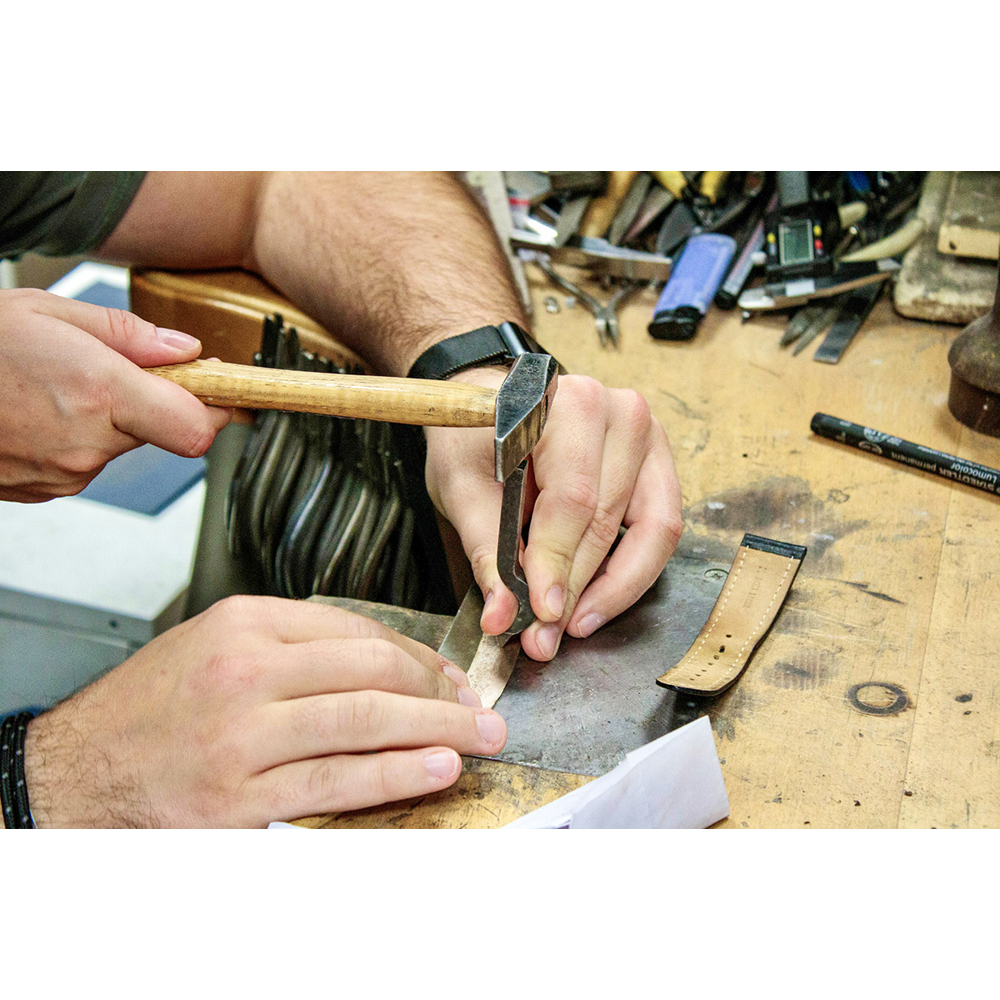 Ein Handwerker formt Metall mit einem kleinen Hammer auf einer Werkbank voller Werkzeuge.