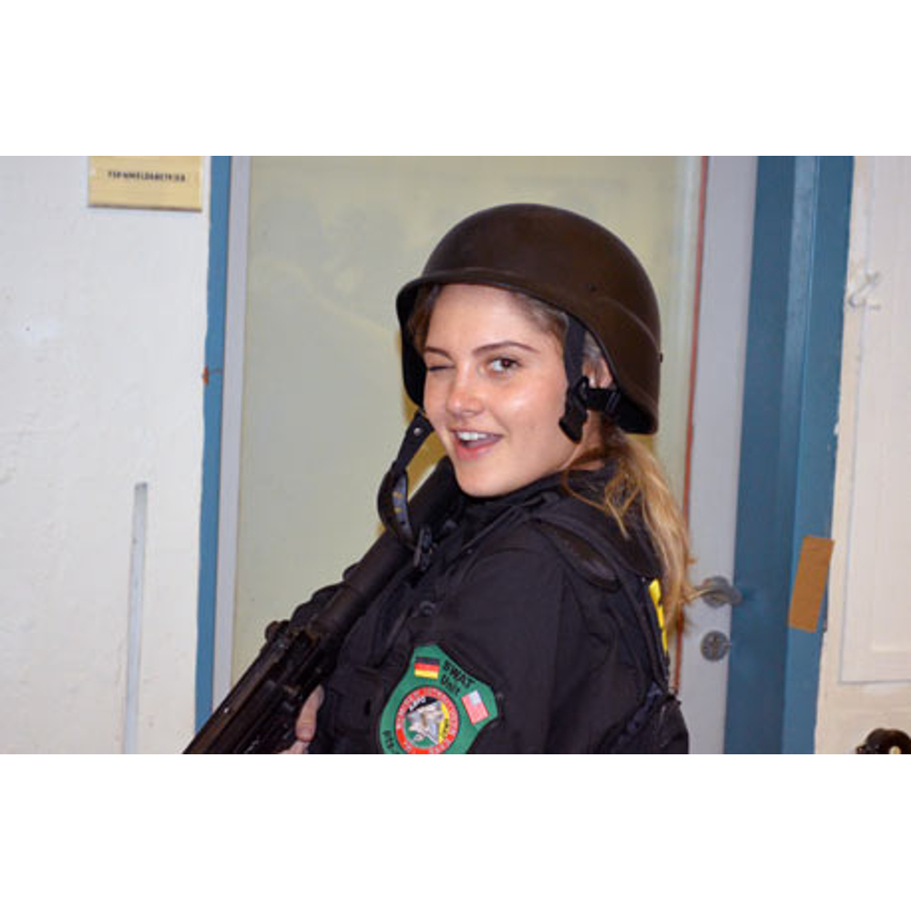 Eine Frau in taktischer Ausrüstung und Helm zwinkert, während sie ein Gewehr hält.