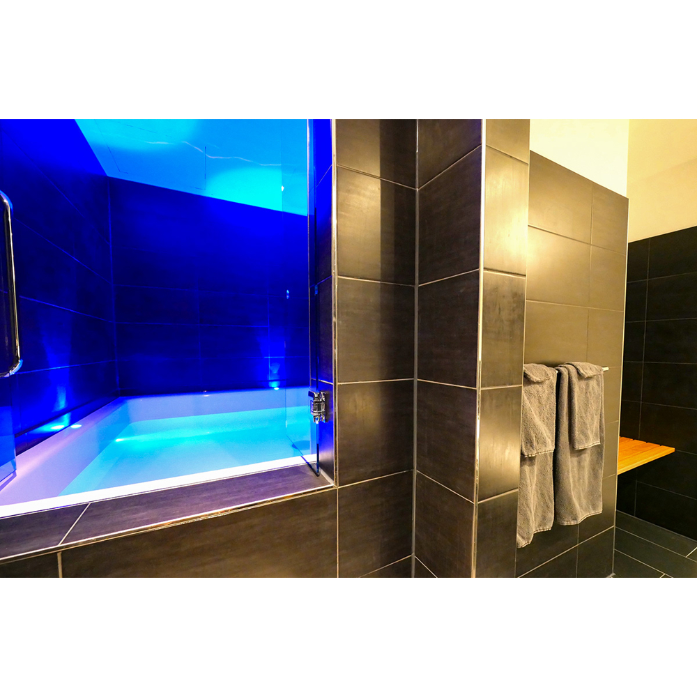 Ein moderner Spa-Raum mit einem blau leuchtenden Sinnesdeprivationstank und grauen Fliesenwänden.