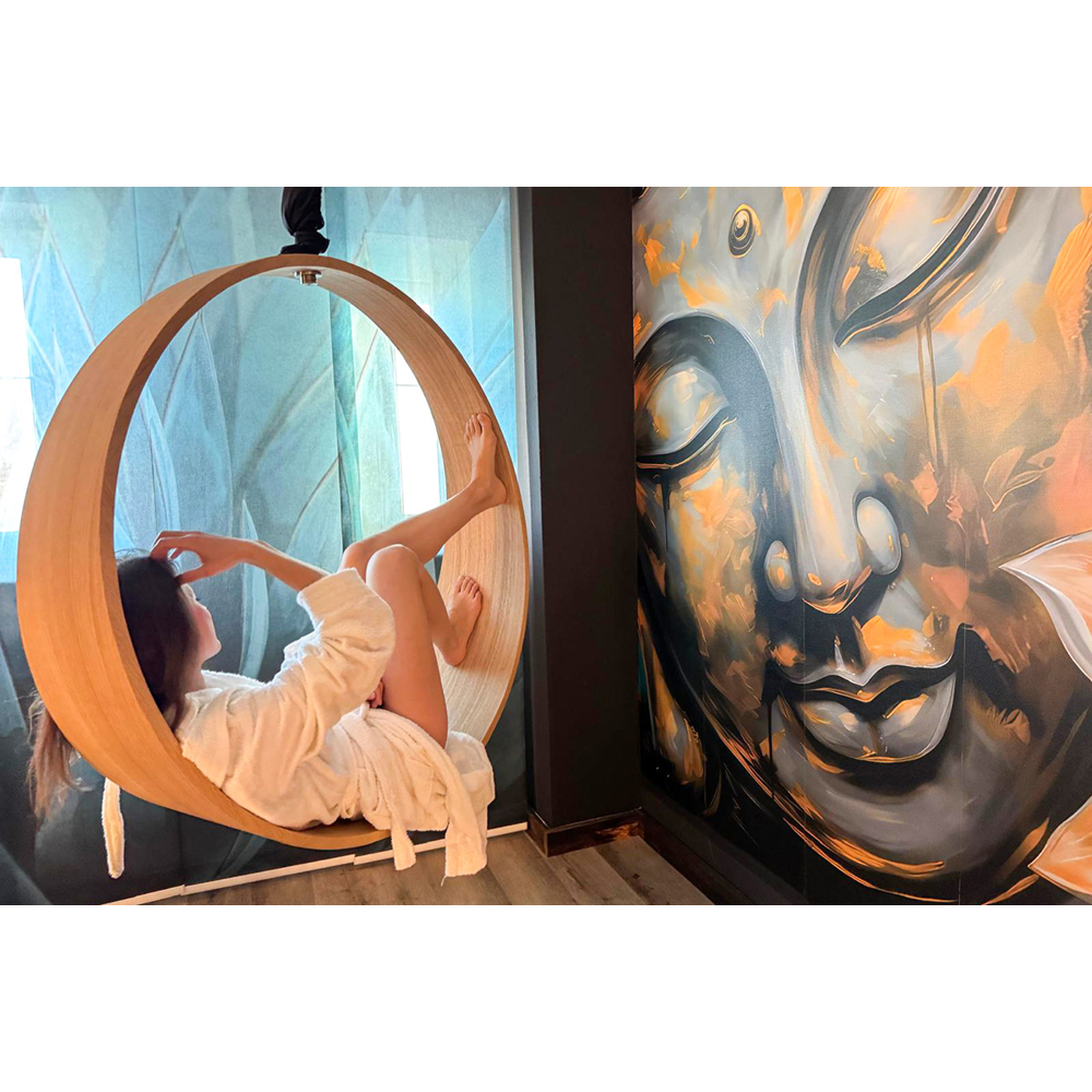 Eine Frau im Bademantel sitzt in einer runden Holzschaukel neben einem großen Buddha-Wandbild.