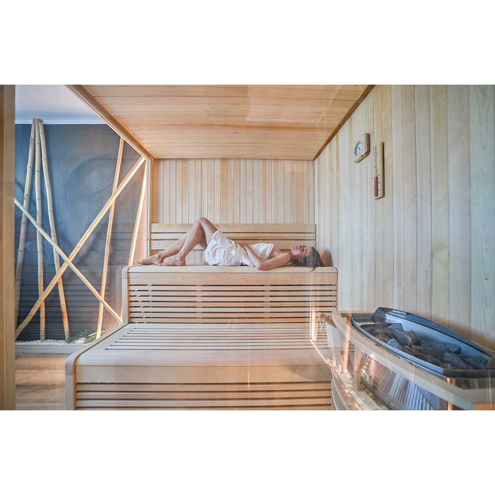 Eine Frau in einem weißen Handtuch entspannt auf einer Holzbank in einer hellen Sauna mit Ofen.