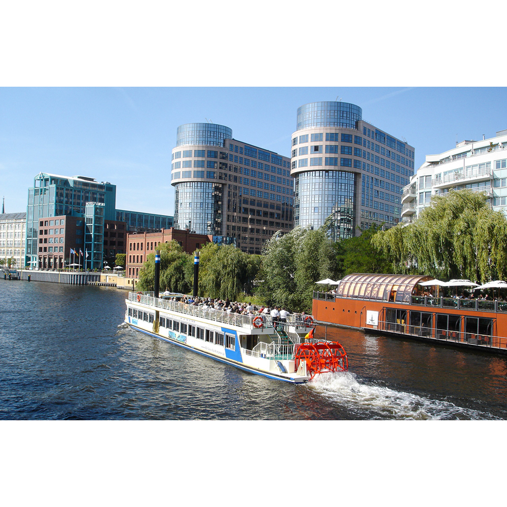 Ein Passagierschiff mit rotem Schaufelrad fährt auf einem Fluss vorbei an modernen Gebäuden und Bäumen.