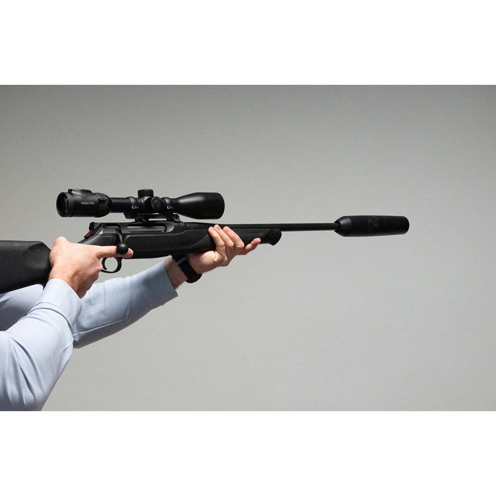 Person hält ein schwarzes Jagdgewehr mit Zielfernrohr und Schalldämpfer vor grauem Hintergrund.