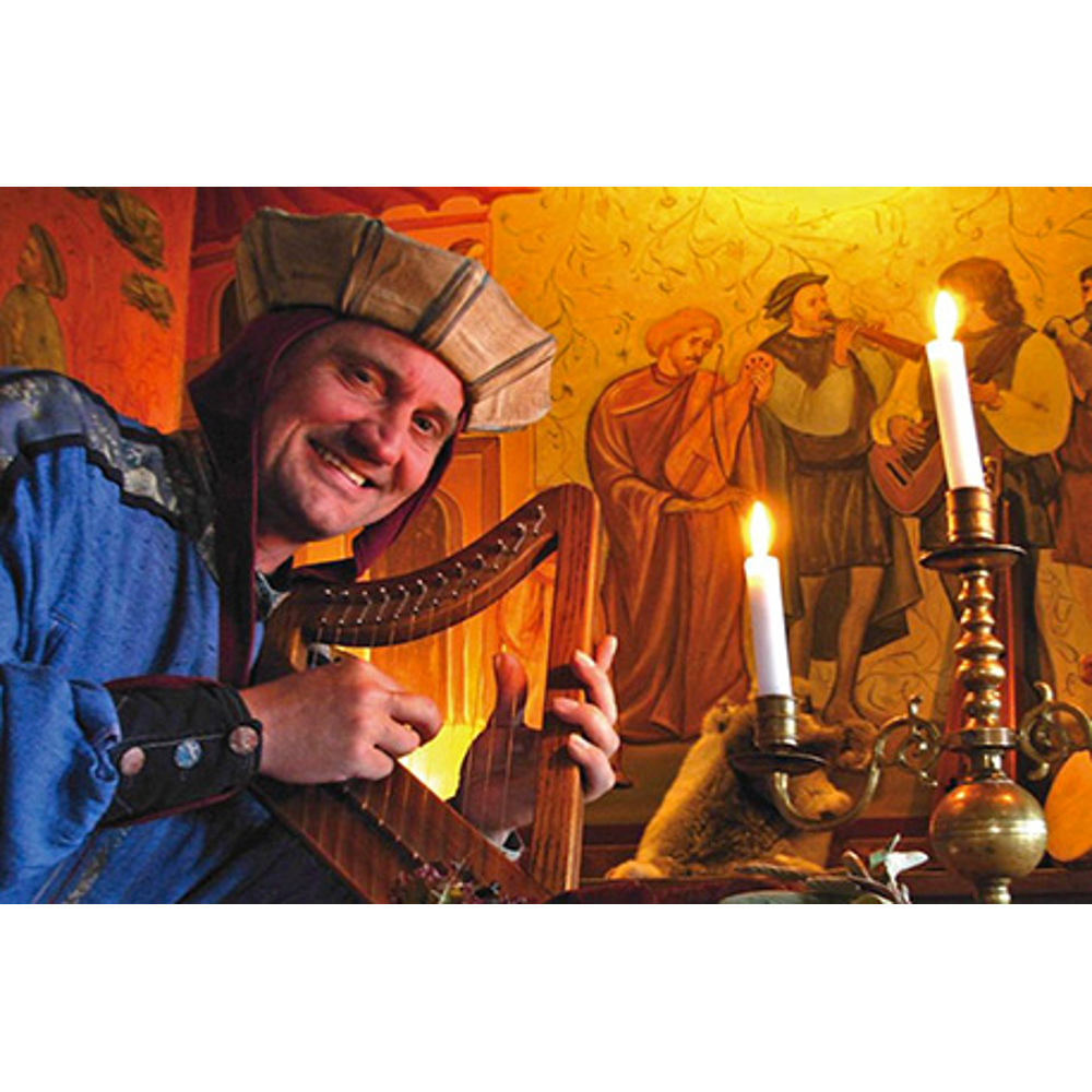Ein mittelalterlich gekleideter Mann lächelt, während er vor einer bemalten Wand eine kleine Harfe spielt.