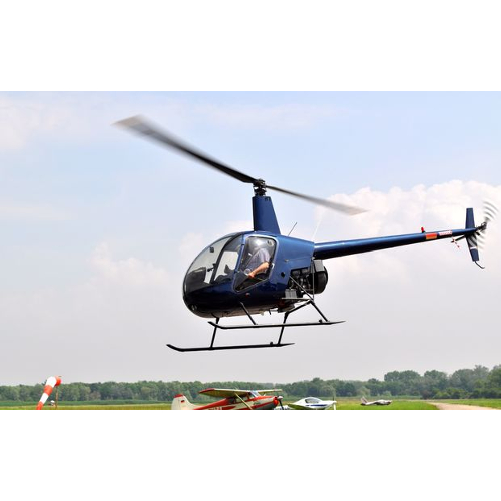 Ein blauer Hubschrauber fliegt über einem grünen Flugplatz, im Hintergrund sind Bäume zu sehen.