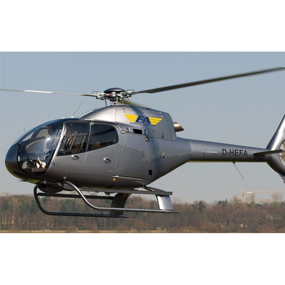 Ein grauer Eurofly-Aviation-Hubschrauber fliegt über einer offenen Landschaft mit Bäumen im Hintergrund.