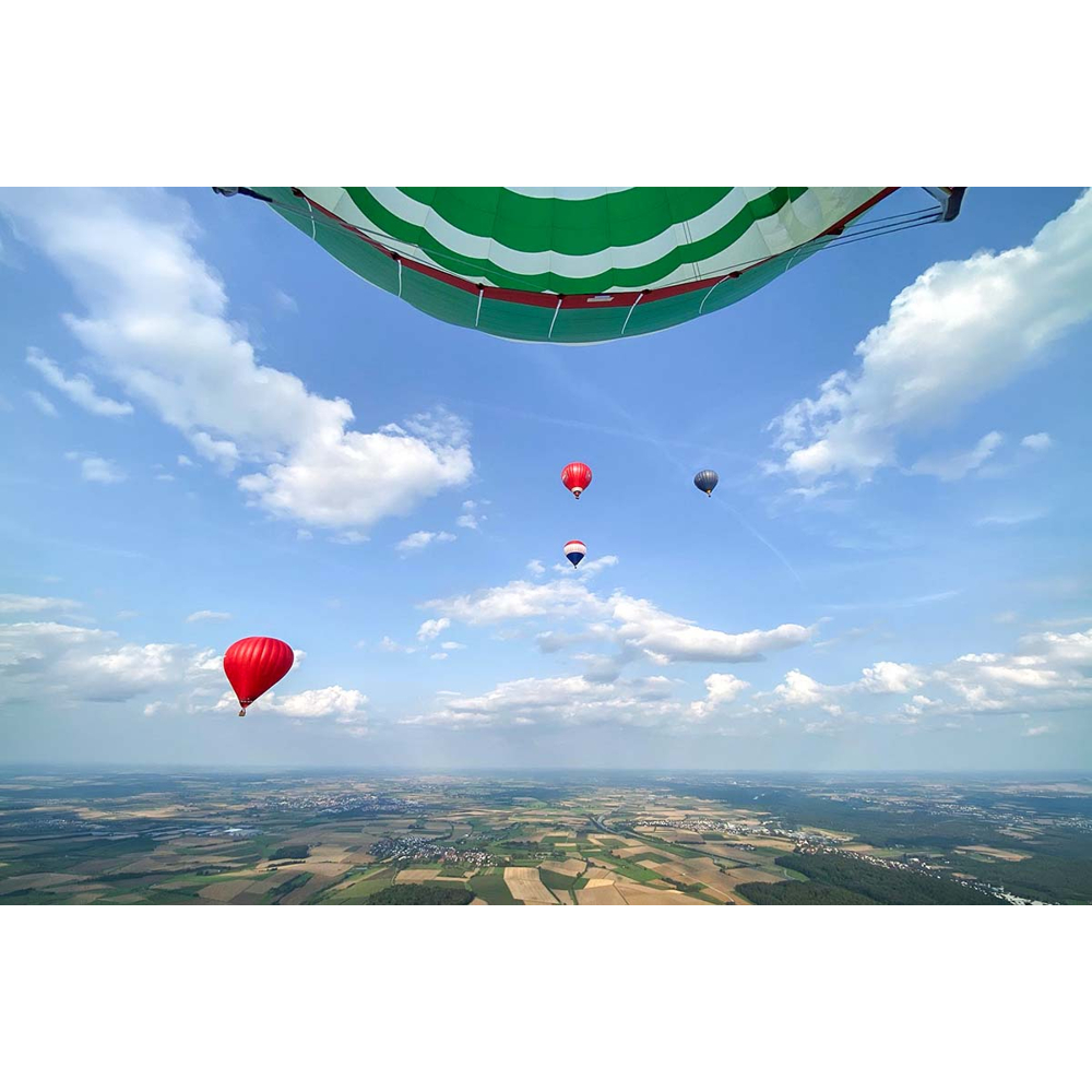 Blick aus einem Heißluftballon auf eine weite Landschaft und andere bunte Ballone am blauen Himmel.