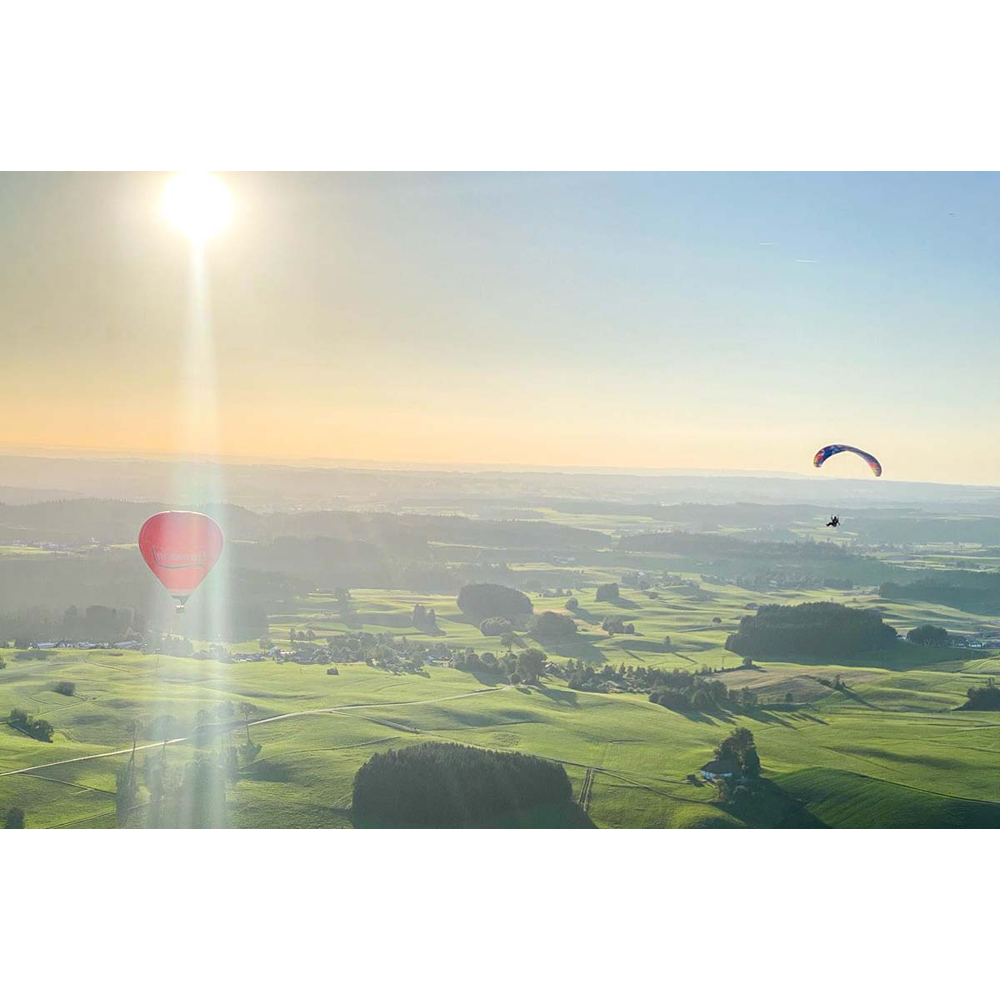 Luftaufnahme einer grünen Landschaft mit Heißluftballon, Gleitschirm und heller Sonne am blauen Himmel.
