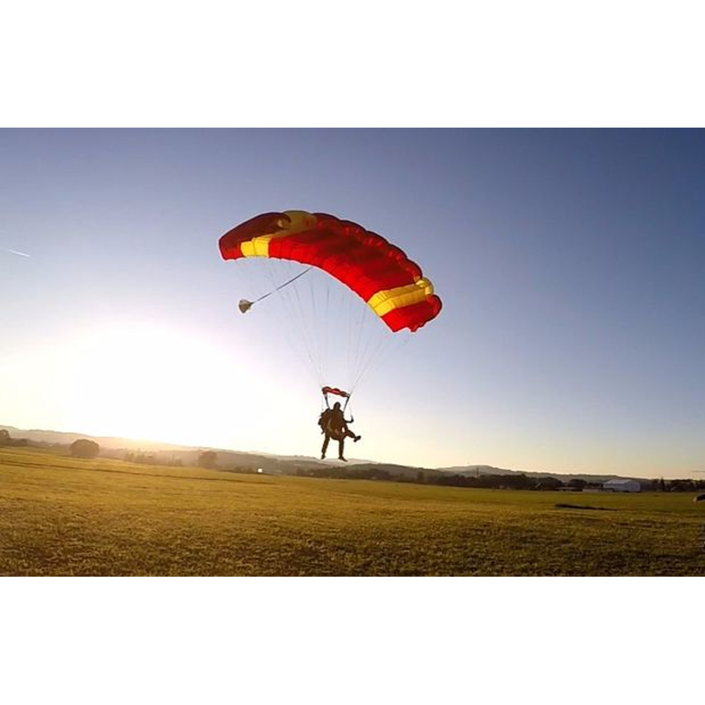 Zwei Fallschirmspringer schweben mit einem rot-gelben Fallschirm bei Sonnenuntergang über ein Feld.