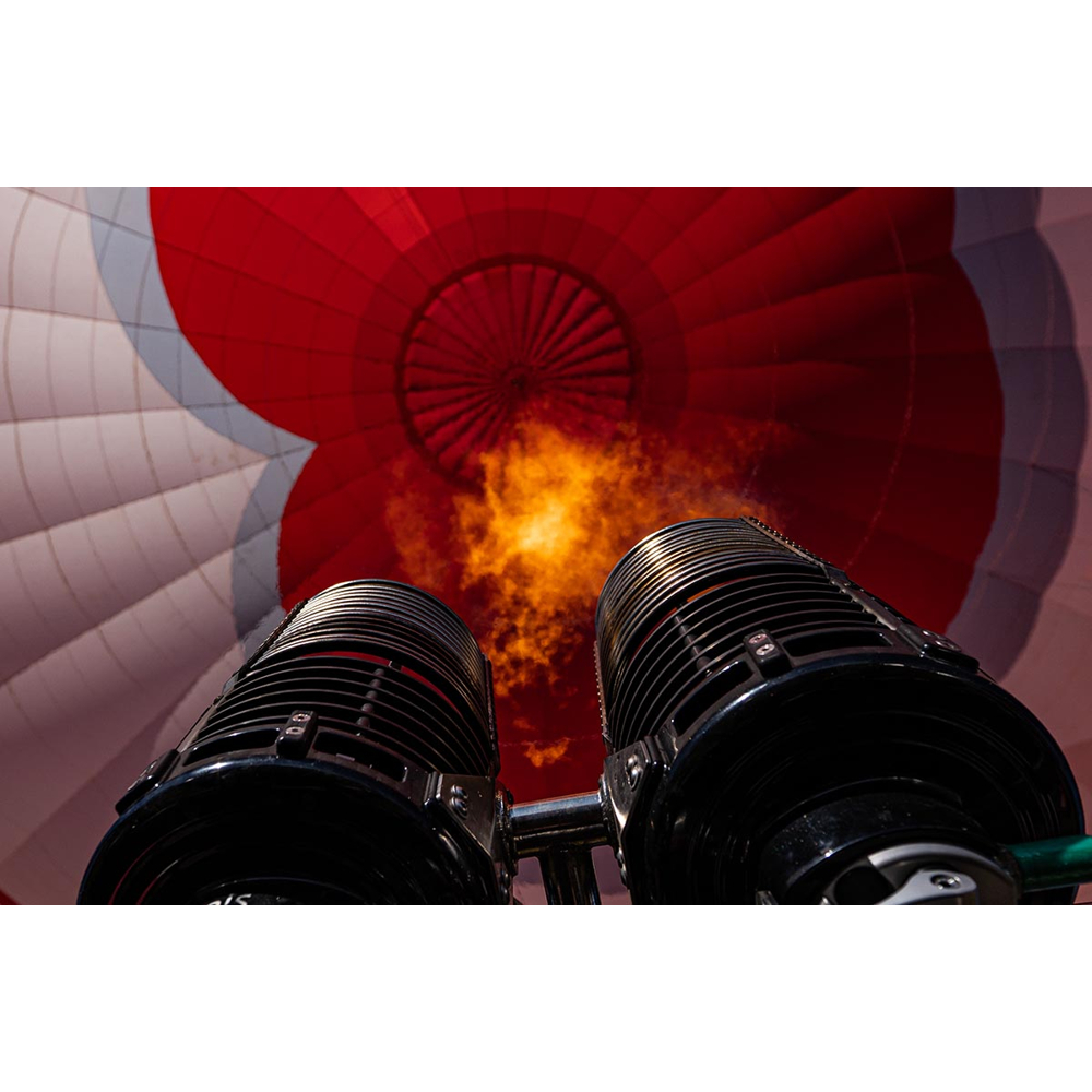 Blick von unten auf zwei Heißluftballonbrenner, die eine helle Flamme in die rote und weiße Ballonhülle richten.