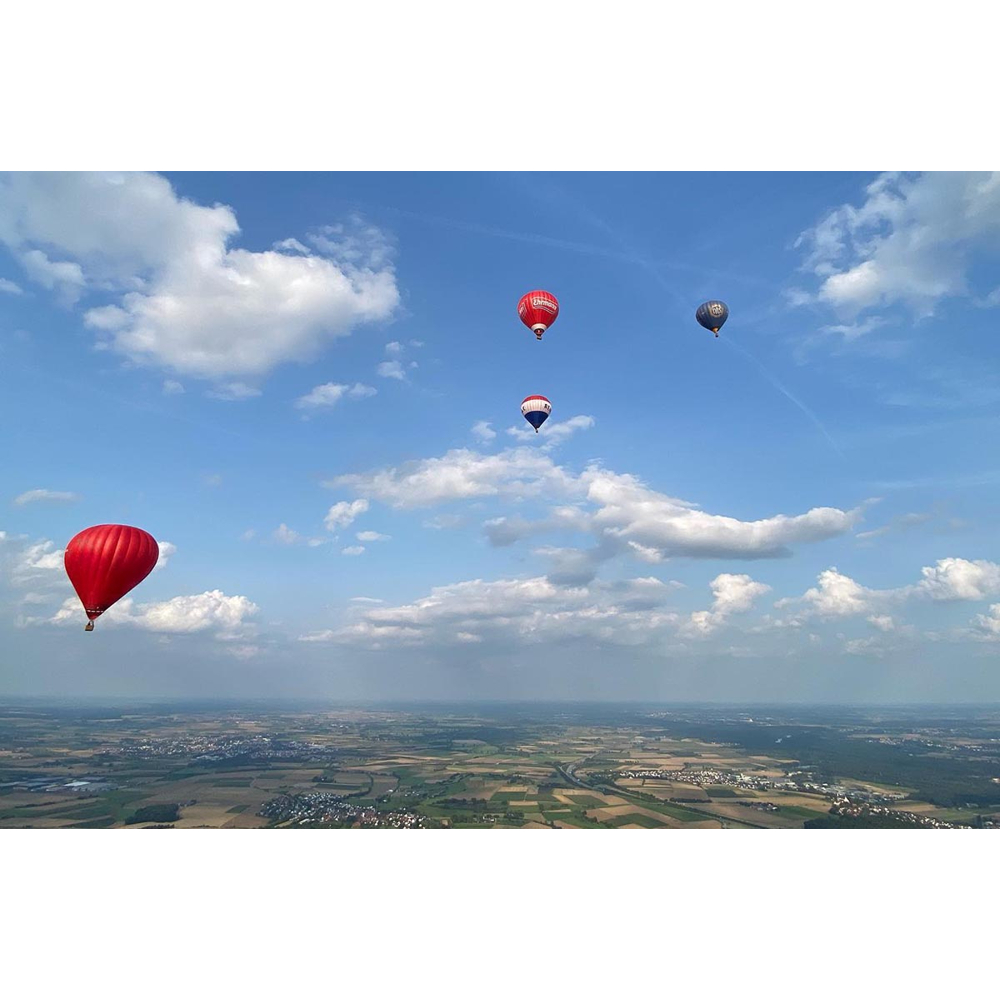 Vier bunte Heißluftballons fliegen am blauen Himmel über einer weiten Landschaft mit Feldern und kleinen Dörfern.