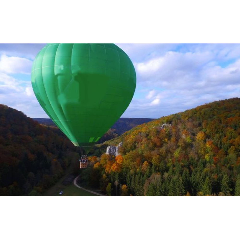 Ein großer grüner Heißluftballon schwebt über herbstlich gefärbten Wäldern und Tälern.