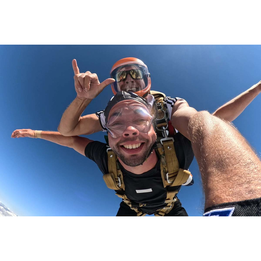 Zwei Männer machen einen Tandemsprung und lächeln in die Kamera vor klarem blauem Himmel.