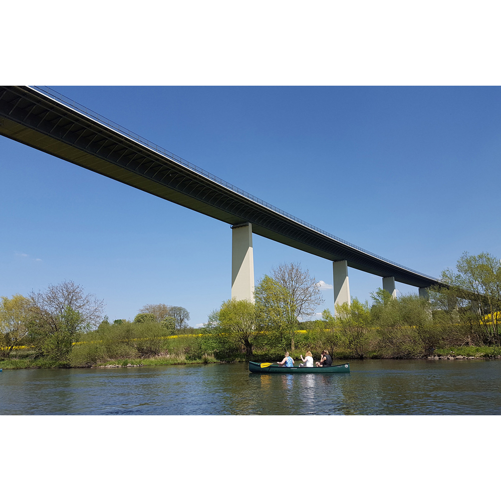 Drei Personen in einem grünen Kanu auf einem Fluss unter einer großen Autobahnbrücke, umgeben von Bäumen und blauem Himmel.