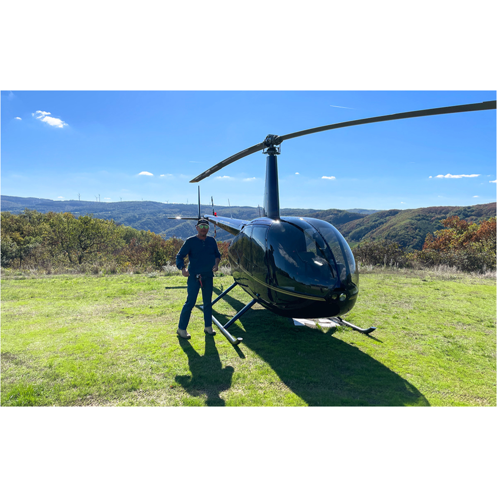 Ein Mann steht neben einem schwarzen Hubschrauber auf einer Wiese vor einer hügeligen Landschaft.