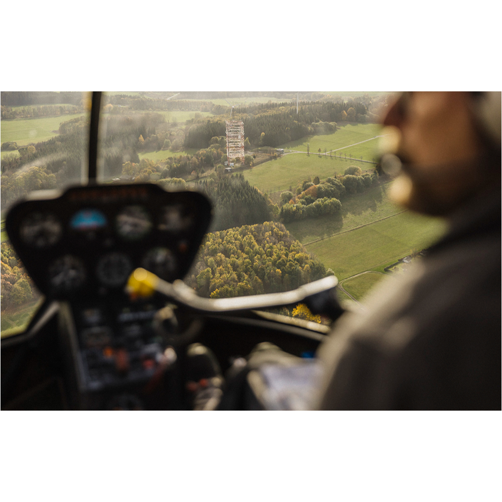 Blick aus einem Hubschrauber-Cockpit auf Armaturen, Pilotenprofil und grüne Landschaft mit einem hohen Turm.