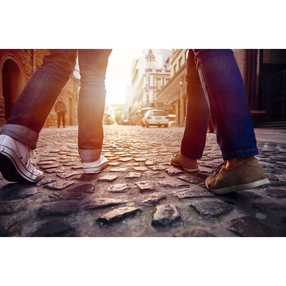 Zwei Personen in Jeans und Schuhen gehen auf einer sonnigen Kopfsteinpflasterstraße in einer Stadt.