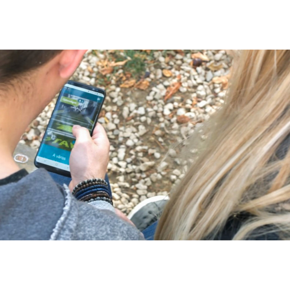 Zwei Personen sitzen im Freien auf Steinen, während eine Person ein Smartphone mit einer grünen App bedient.