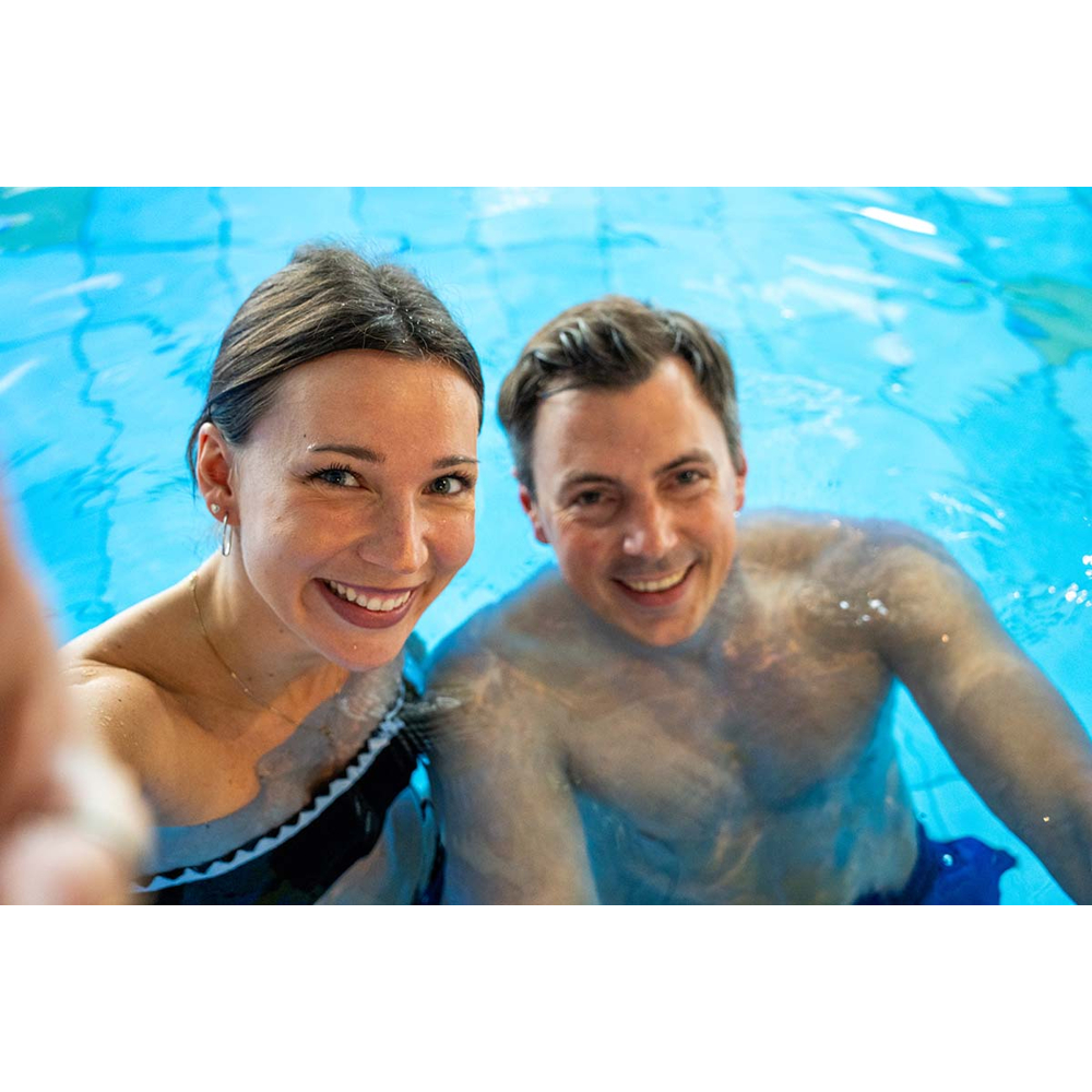 Ein lächelndes Paar macht zusammen ein Selfie beim Schwimmen in einem blauen Pool.