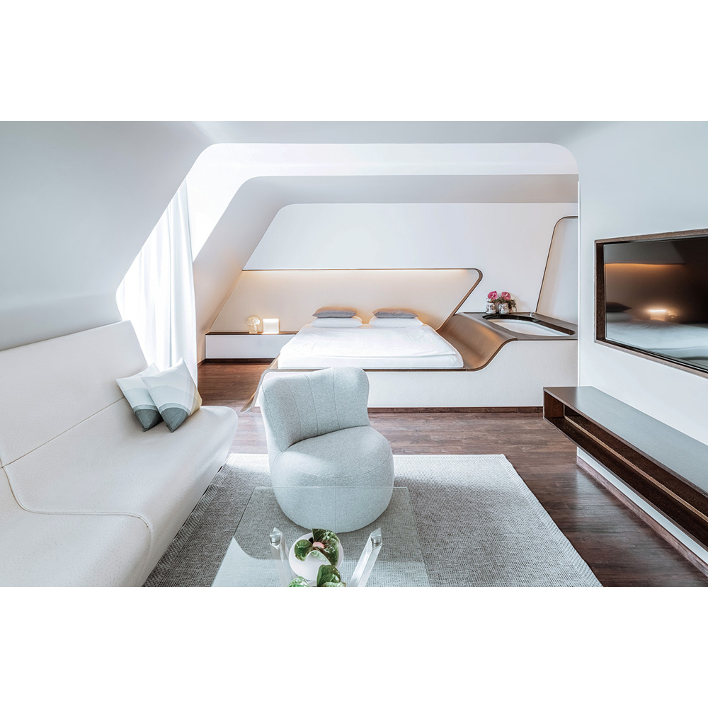 Ein modernes Hotelzimmer mit weißem Sofa, rundem Sessel, Bett, Badewanne und an der Wand montiertem Fernseher.