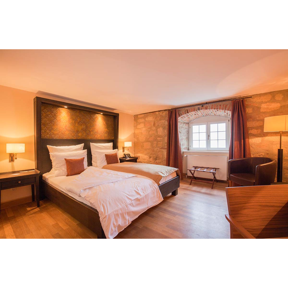 Gemütliches Schlafzimmer mit großem Doppelbett, Steinwand, Fenstern mit roten Vorhängen und Sesseln auf Holzboden.