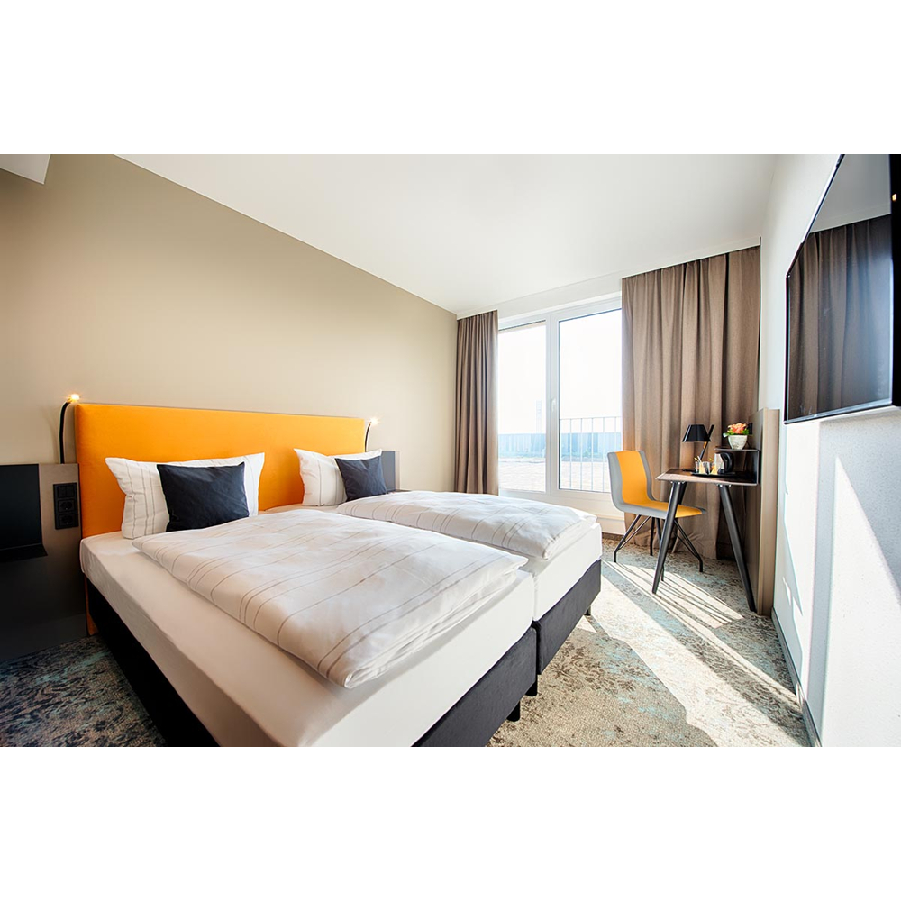 Moderne Hotelzimmer mit zwei Betten, orangem Kopfkissen, Schreibtisch und großem Fenster mit Vorhängen.