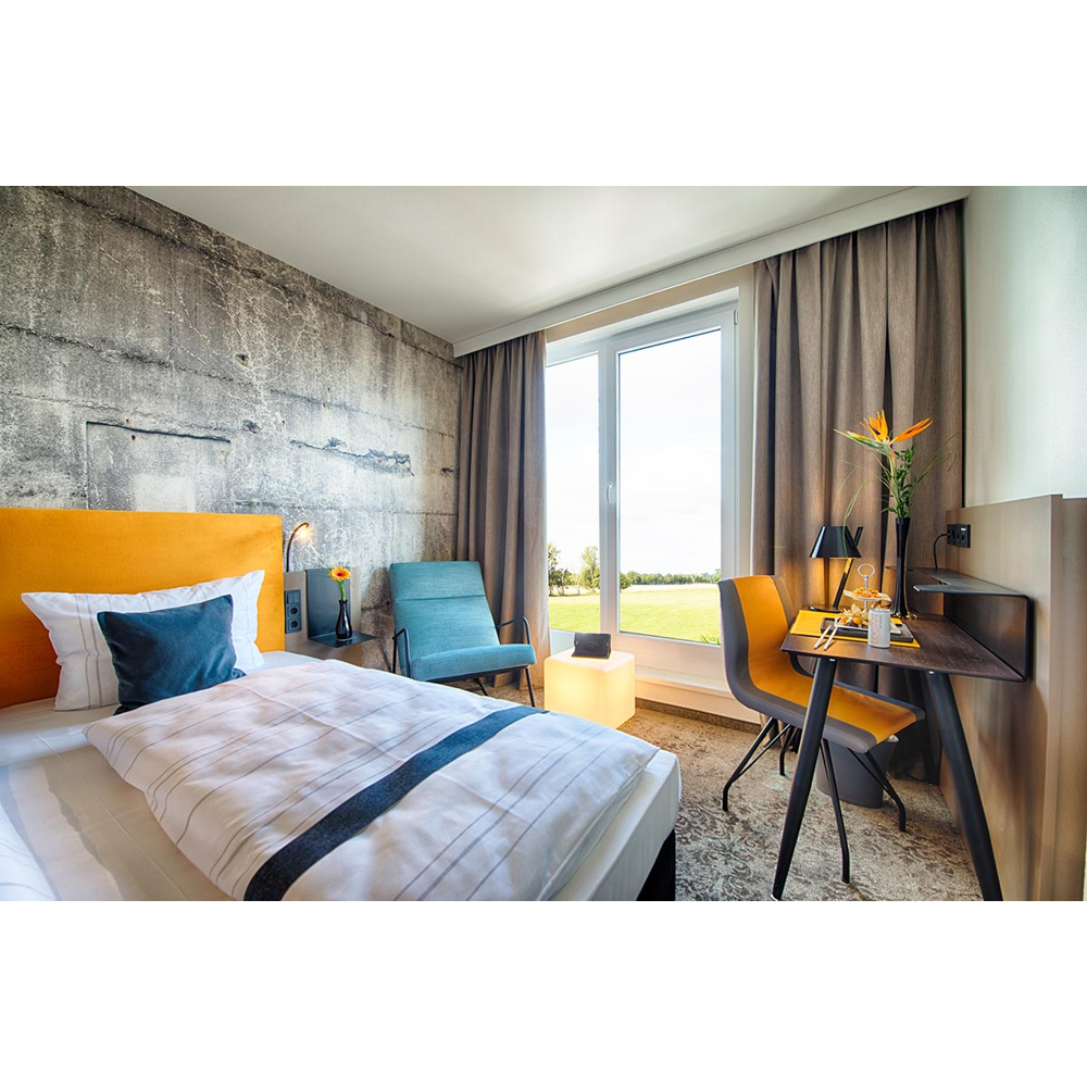 Ein modernes Hotelzimmer mit gelbem Bett, Betonwand, Schreibtisch und Fenster mit Blick ins Grüne.