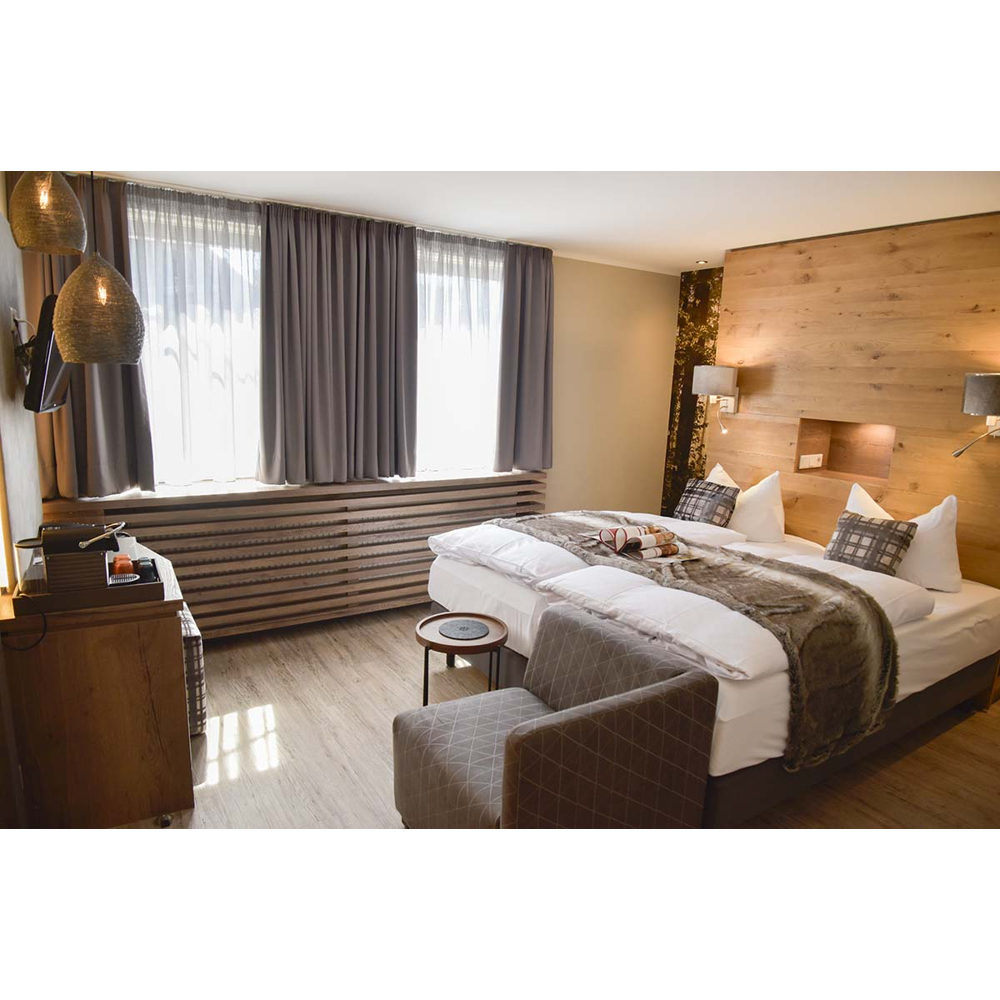 Gemütliches Hotelzimmer mit großem Bett, Holzpaneel-Wand, Sesseln und Fenstern mit Vorhängen.