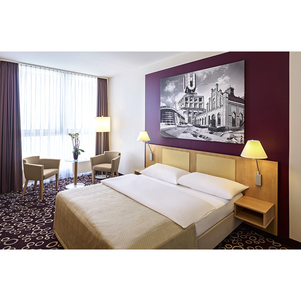 Moderne Hotelzimmer mit großem Bett, dunkler Wand, großem Schwarz-Weiß-Bild und Sitzbereich am Fenster mit Mustervorhängen.