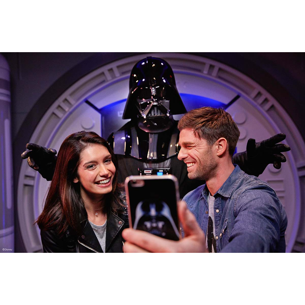 Ein lächelndes Paar macht ein Selfie mit Darth Vader, der hinter ihnen in einer Sci-Fi-Kulisse steht.